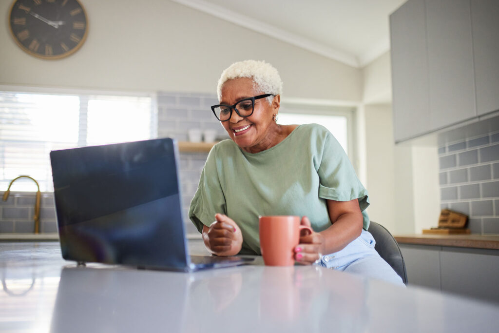 An elderly woman with short white hair and glasses smiles while looking at her laptop in a bright kitchen. She holds a peach-colored mug.