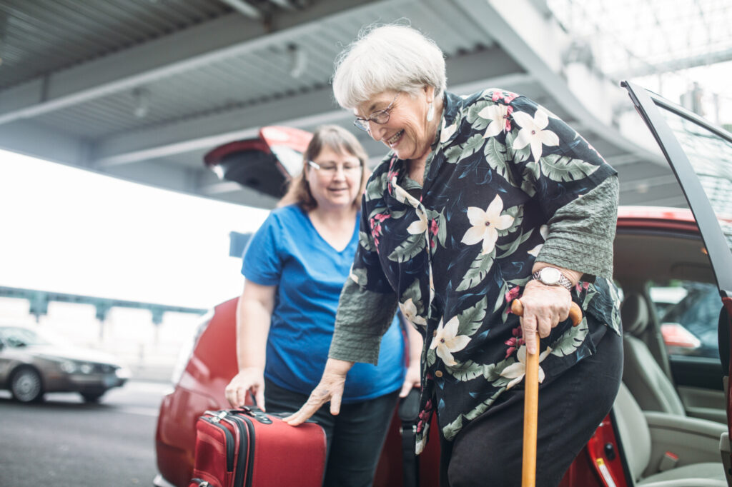 A mature woman in her 70's gets out of a car while holding a cane with a female friend at the airport. The friend hands the woman her luggage. Both women have a cheerful smile on their faces.