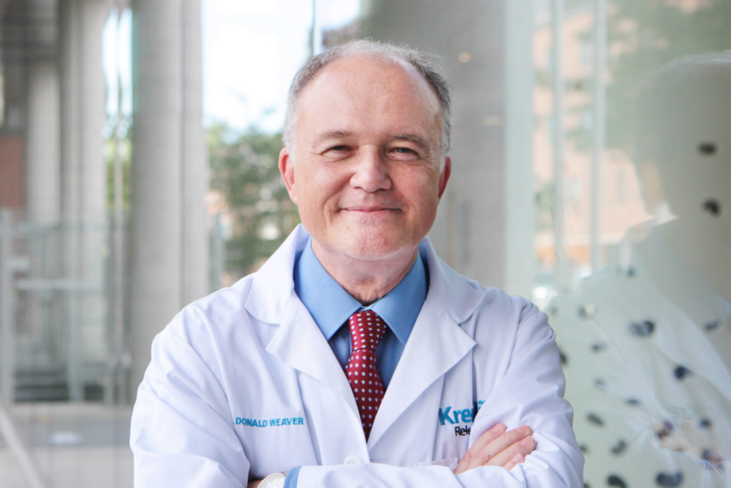 Dr. Donald Weaver smiles with his arms crossed while wearing a white lab coat over a blue collared buttoned shirt and a maroon coloured tie.